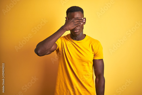 Young african american man wearing casual t-shirt standing over isolated yellow background covering eyes with hand, looking serious and sad. Sightless, hiding and rejection concept