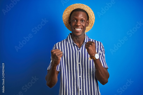 African american man wearing striped shirt and summer hat over isolated blue background celebrating surprised and amazed for success with arms raised and open eyes. Winner concept. © Krakenimages.com