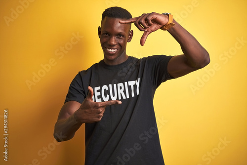 African american safeguard man wearing security uniform over isolated yellow background smiling making frame with hands and fingers with happy face. Creativity and photography concept.