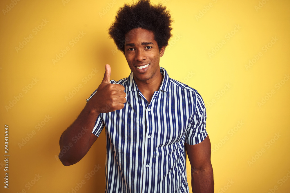 American man with afro hair wearing striped shirt standing over isolated yellow background doing happy thumbs up gesture with hand. Approving expression looking at the camera with showing success. American man with afro hair wearing striped shirt standing over isolated yellow background doing happy thumbs up gesture with hand. Approving expression looking at the camera with showing success.