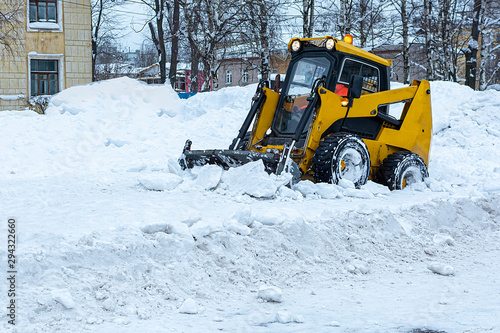 A small yellow tractor removes snow on the street. The concept of snow removal after heavy snow falls and blizzards, problems associated with weather conditions.