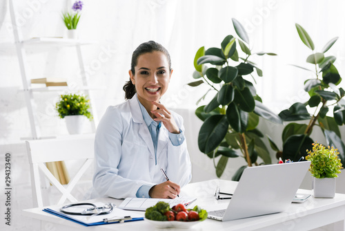 happy nutritionist in white coat near vegetables and laptop photo