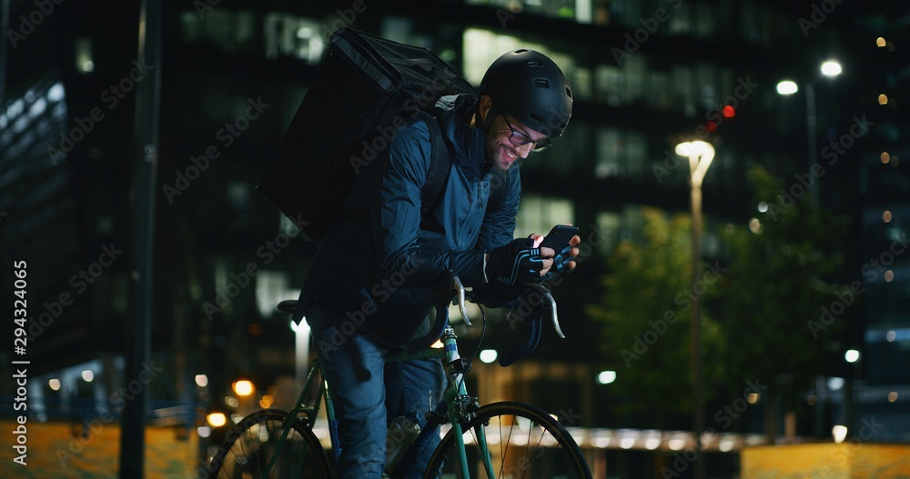 Portrait of an young delivery courier with bicycle is controlling a customer addresses by using smartphone with gps in the evening in a city center.