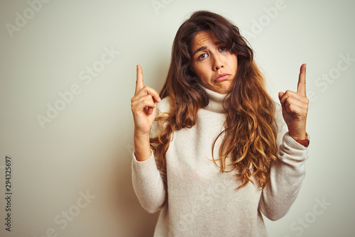 Young beautiful woman wearing winter sweater standing over white isolated background Pointing up looking sad and upset, indicating direction with fingers, unhappy and depressed.
