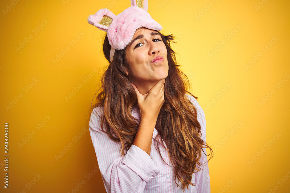 Young woman wearing pajama and sleep mask standing over yellow isolated background Touching painful neck, sore throat for flu, clod and infection Young woman wearing pajama and sleep mask standing over yellow isolated background Touching painful neck, sore throat for flu, clod and infection