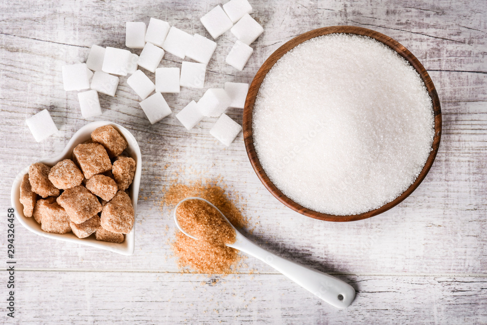 White fresh sugar on white table. Sugar in wood bowl. Brown sugar in heart bowl with spoon and brown cube. White fresh sugar on white table. Sugar in wood bowl. Brown sugar in heart bowl with spoon and brown cube.