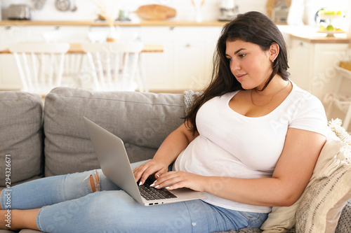 Cute teenage girl with extra pounds keeping blog, typing new post on her account, chatting with her followers online, having happy facial expression, sitting on couch with portable computer