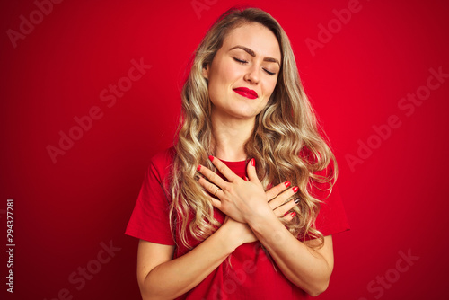 Young beautiful woman wearing basic t-shirt standing over red isolated background smiling with hands on chest with closed eyes and grateful gesture on face. Health concept.