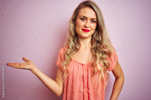 Young beautiful woman wearing t-shirt standing over pink isolated background smiling cheerful presenting and pointing with palm of hand looking at the camera.