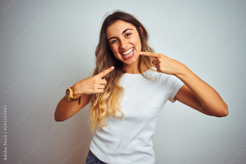 Young beautiful woman wearing casual white t-shirt over isolated background smiling cheerful showing and pointing with fingers teeth and mouth. Dental health concept. Young beautiful woman wearing casual white t-shirt over isolated background smiling cheerful showing and pointing with fingers teeth and mouth. Dental health concept.