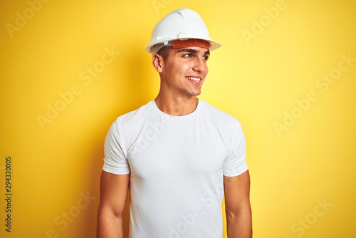 Young handsome man wearing construction helmet over yellow isolated background looking away to side with smile on face  natural expression. Laughing confident.