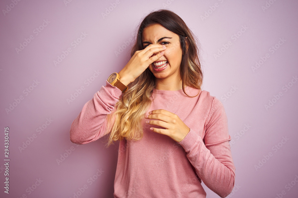 Young beautiful woman wearing a sweater over pink isolated background smelling something stinky and disgusting, intolerable smell, holding breath with fingers on nose. Bad smells concept. Young beautiful woman wearing a sweater over pink isolated background smelling something stinky and disgusting, intolerable smell, holding breath with fingers on nose. Bad smells concept.