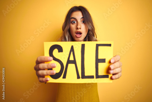 Young beautiful woman holding sale poster over yellow isolated background scared in shock with a surprise face, afraid and excited with fear expression