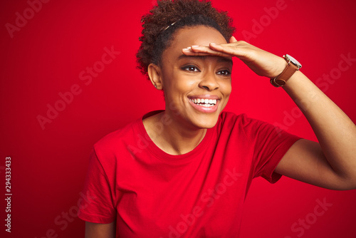 Young beautiful african american woman with afro hair over isolated red background very happy and smiling looking far away with hand over head. Searching concept.