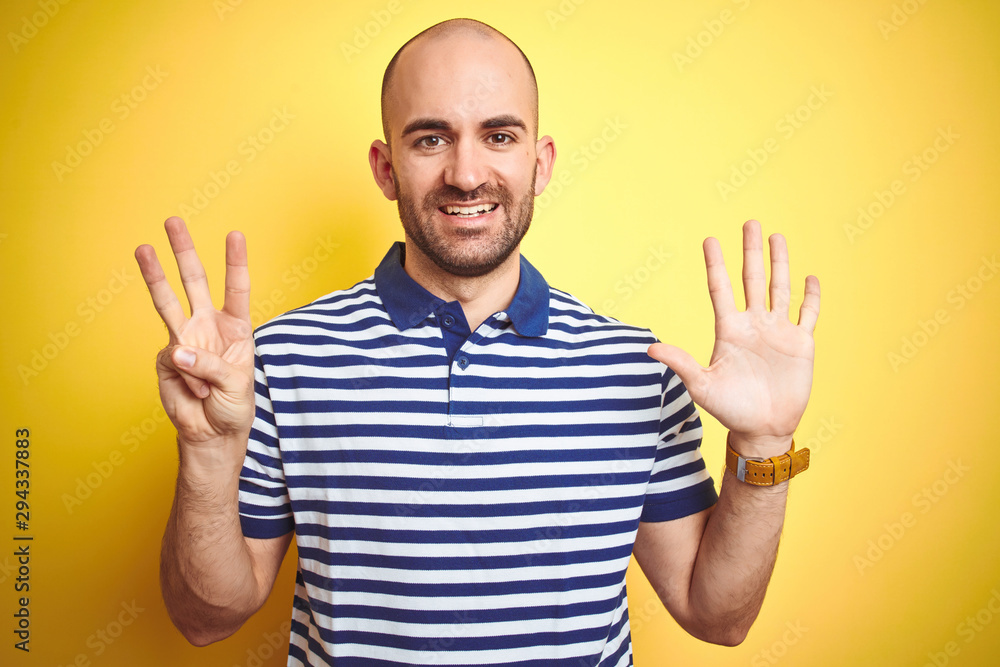 Naklejka premium Young bald man with beard wearing casual striped blue t-shirt over yellow isolated background showing and pointing up with fingers number eight while smiling confident and happy.