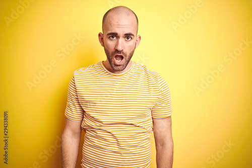 Young bald man with beard wearing casual striped t-shirt over yellow isolated background In shock face, looking skeptical and sarcastic, surprised with open mouth
