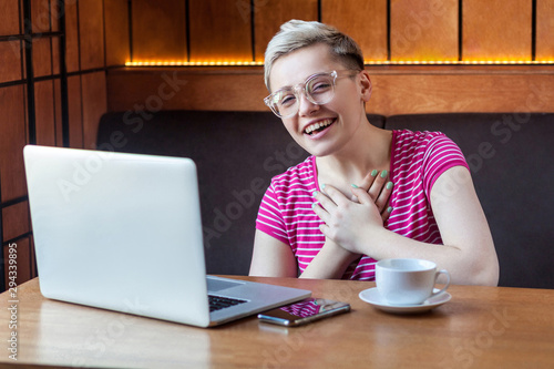 Portrait of happy smile young adult woman with short hair in pink t-shirt and eyeglasses is sitting with laptop in cafe and laughfing, looking at camera with toothy smile, indoor, lifestyle photo