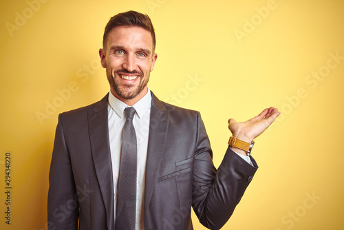 Young handsome business man over yellow isolated background smiling cheerful presenting and pointing with palm of hand looking at the camera.