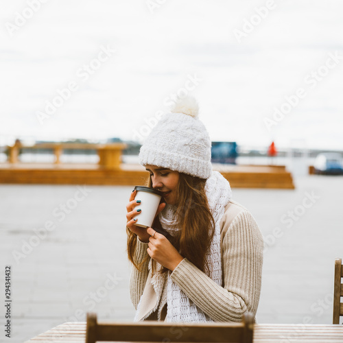 Beautiful young girl drinking coffee, tea from plastic mug in autumn, winter. A woman with long hair in warm clothes sitting on waterfront on Baltic sea in port, warmed by hot drink