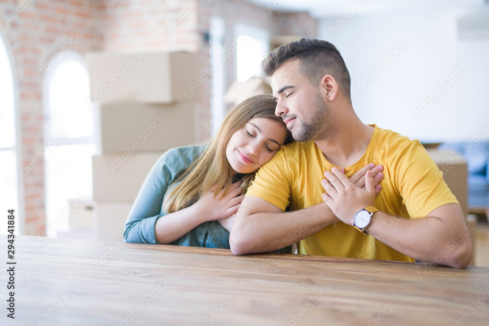 Young couple sitting on the table movinto to new home with carboard boxes behind them smiling with hands on chest with closed eyes and grateful gesture on face. Health concept. Young couple sitting on the table movinto to new home with carboard boxes behind them smiling with hands on chest with closed eyes and grateful gesture on face. Health concept.