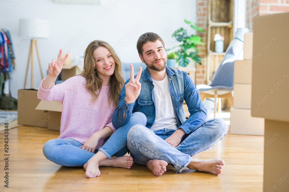 Young beautiful couple moving to a new house sitting on the floor smiling with happy face winking at the camera doing victory sign. Number two. Young beautiful couple moving to a new house sitting on the floor smiling with happy face winking at the camera doing victory sign. Number two.