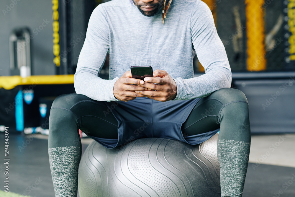 Cropped image of young sportsman sitting on big fitness ball and checking health application on smartphone Cropped image of young sportsman sitting on big fitness ball and checking health application on smartphone