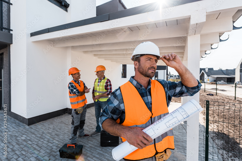 selective focus of constructor with blueprint touching helmet near men selective focus of constructor with blueprint touching helmet near men