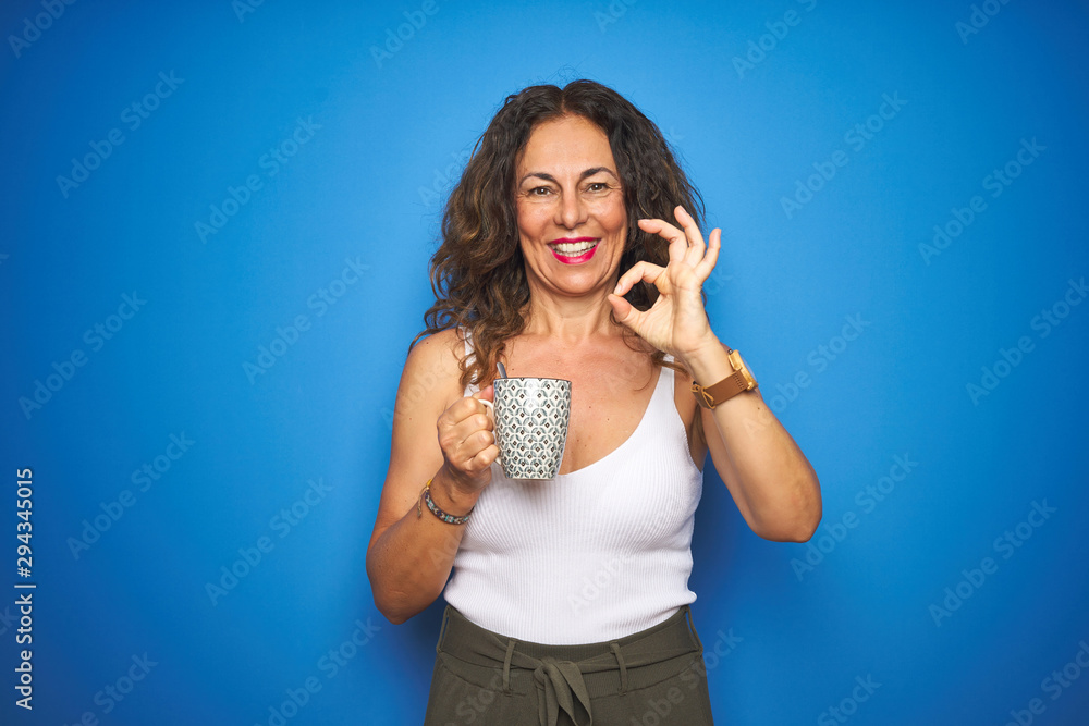 Middle age senior woman drinking a cup of coffee over blue isolated background doing ok sign with fingers, excellent symbol Middle age senior woman drinking a cup of coffee over blue isolated background doing ok sign with fingers, excellent symbol