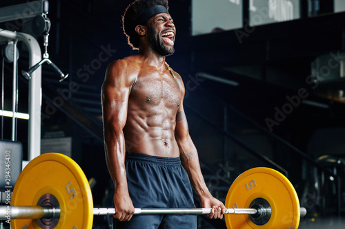 Shirtless strong young sportsman shouting when lifting heavy weight in gym