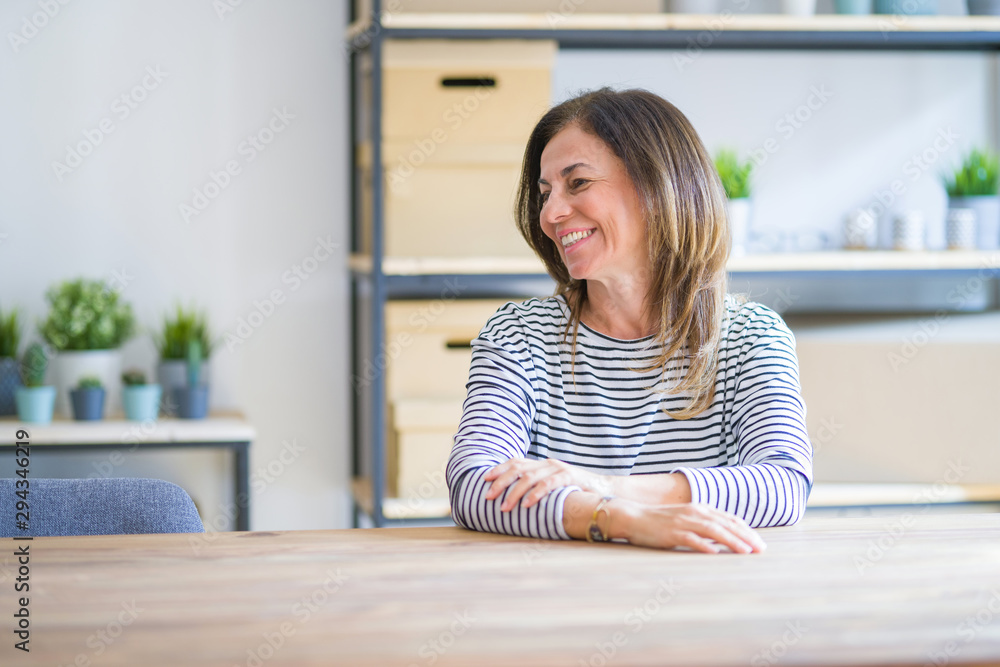 Middle age senior woman sitting at the table at home looking away to side with smile on face, natural expression. Laughing confident. Middle age senior woman sitting at the table at home looking away to side with smile on face, natural expression. Laughing confident.