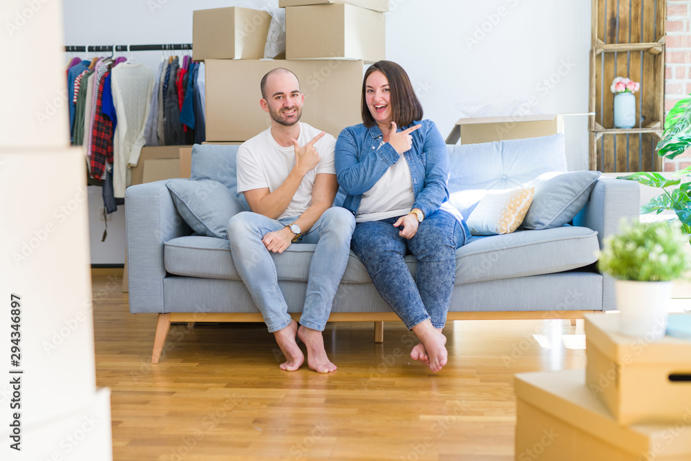 Young couple sitting on the sofa arround cardboard boxes moving to a new house cheerful with a smile of face pointing with hand and finger up to the side with happy and natural expression on face