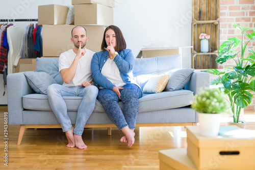 Young couple sitting on the sofa arround cardboard boxes moving to a new house asking to be quiet with finger on lips. Silence and secret concept. © Krakenimages.com