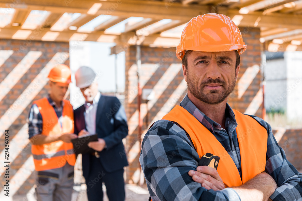 selective focus of handsome man in helmet standing with crossed arms near coworker and businessman selective focus of handsome man in helmet standing with crossed arms near coworker and businessman