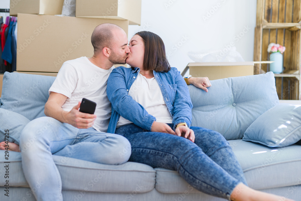 Young couple sitting on the sofa at new home using smartphone, hugging and smiling happy for moving to a new apartment
