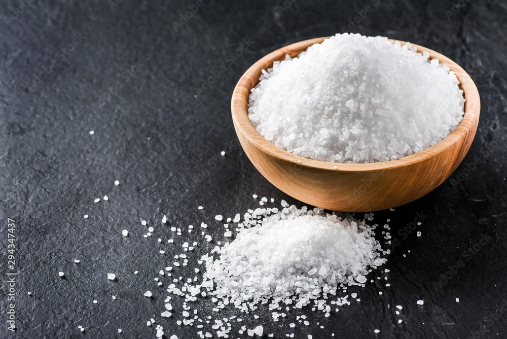 Salt in wooden olive bowl on stone table. Salt top view. Sea salt on black background. Salt in wooden olive bowl on stone table. Salt top view. Sea salt on black background.