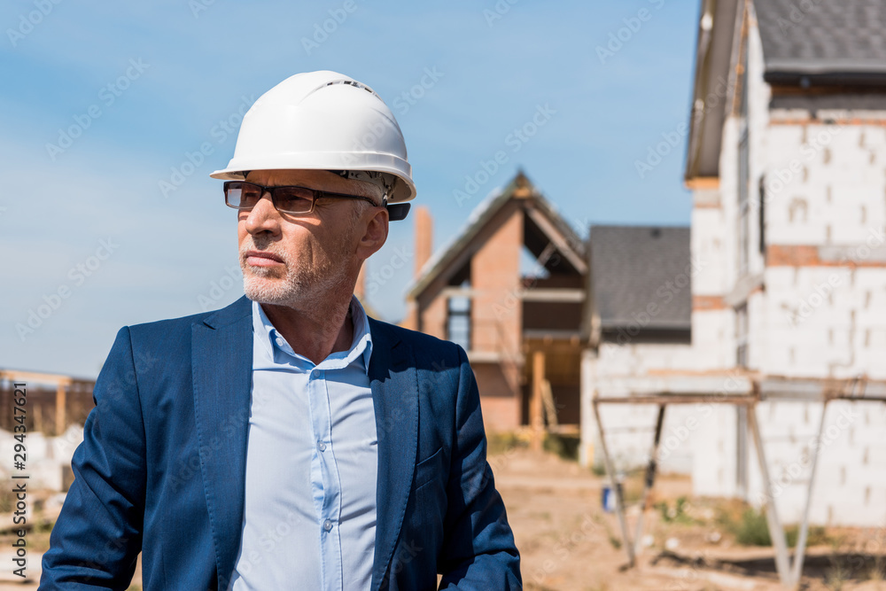 mature and bearded businessman in suit and helmet near buildings mature and bearded businessman in suit and helmet near buildings