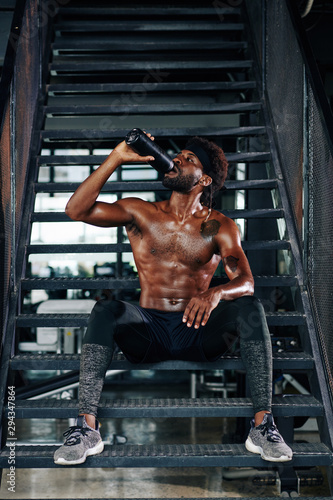 Fit shirtless Black man sitting on metal steps and drinking fresh water from the bottle after working out