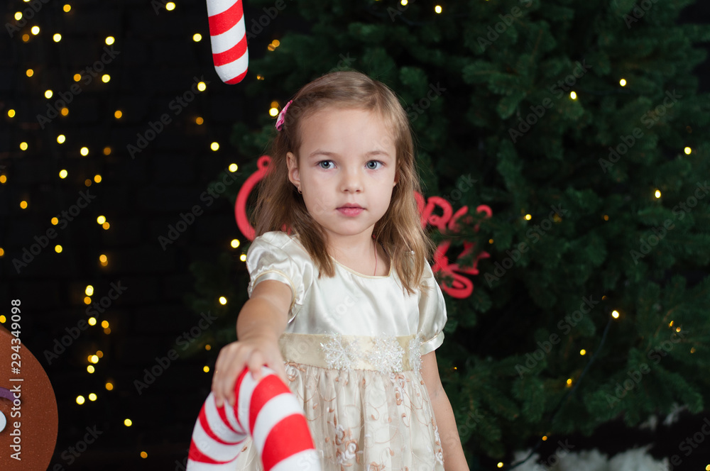 Little beautiful laughing girl in a beige dress near the Christmas tree plays with a big candy, smiling