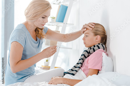 mother holding digital thermometer and touching forehead of daughter