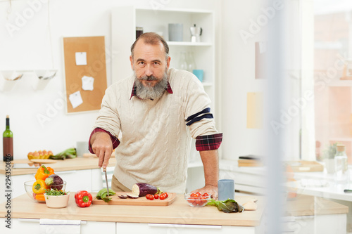 Waist up portrait of bearded senior man cutting vegetables for healthy salad and looking at camera while posing in kitchen, copy space