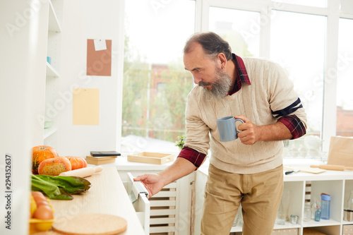Side view portrait of bearded senior man opening kitchen cupboards while looking for breakfast in morning, copy space