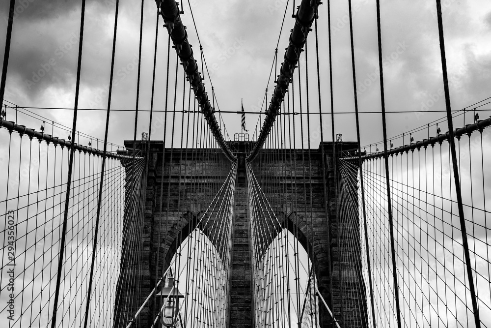 Fototapeta premium Brookyln Bridge Nowy Jork Manhattan czarno-biała grafika w skali szarości niebo chmury inżynier budowlany East River widok punkt orientacyjny kable podwieszane statyka łuki filary pilon murowane symetria