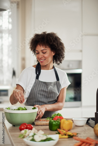 Attractive mixed race woman in apron standing in kitchen and putting chopped lettuce in bowl. Meal preparation concept.