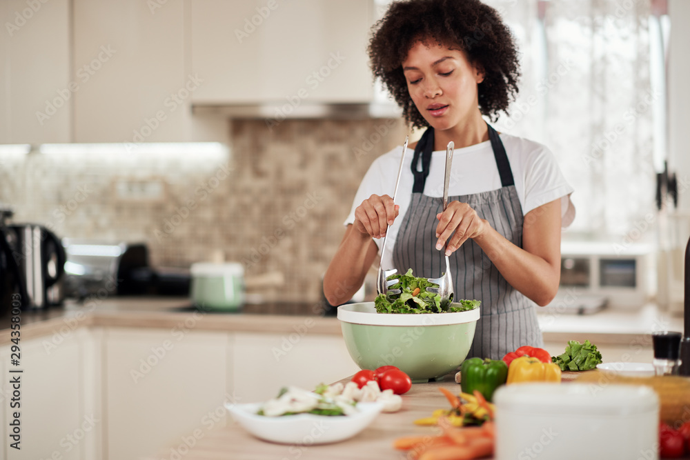 Attractive mixed race woman with curly hair and in apron mixing salad while standing in domestic kitchen.