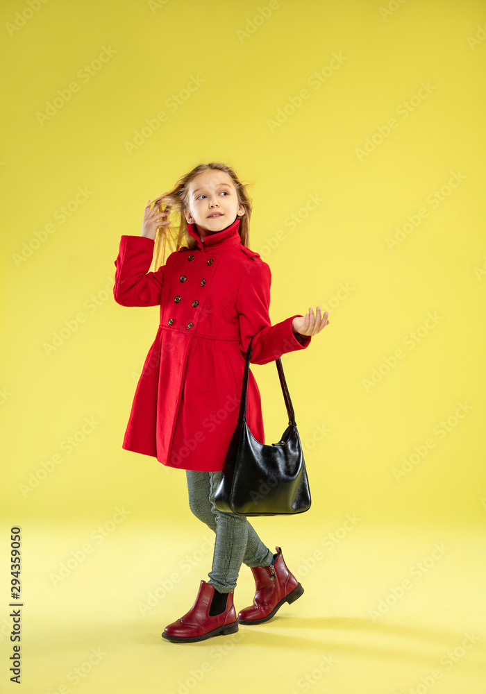 A full length portrait of a bright fashionable girl in a red raincoat with black bag posing on yellow studio background. Looks happy. Autumn and spring fashion for kids. Cute stylish blonde girl. A full length portrait of a bright fashionable girl in a red raincoat with black bag posing on yellow studio background. Looks happy. Autumn and spring fashion for kids. Cute stylish blonde girl.