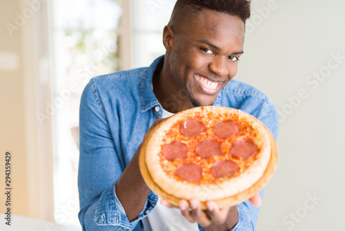 Handsome young african man holding and showing smiling proud homemade pepperoni pizza