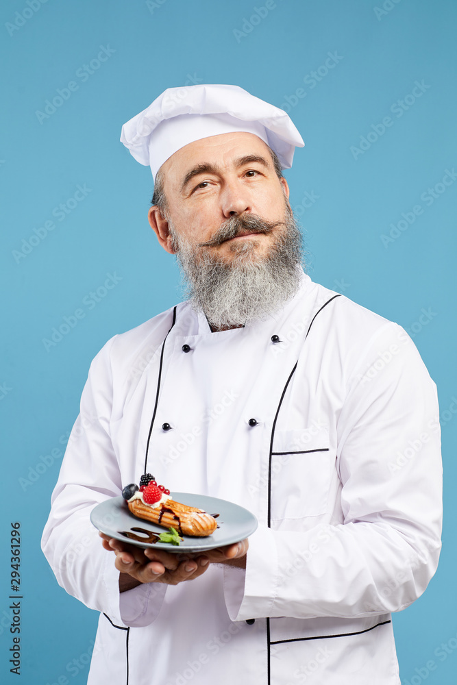 Waist up portrait of cheerful senior chef presenting beautiful dessert while standing against blue background Waist up portrait of cheerful senior chef presenting beautiful dessert while standing against blue background