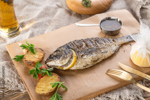 grilled dorado fish with lemon and potato wedges on wooden board and glass of beer on wooden table