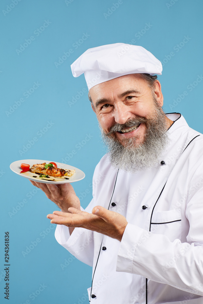 Portrait of cheerful senior chef presenting Italian dish while standing against blue background and smiling at camera Portrait of cheerful senior chef presenting Italian dish while standing against blue background and smiling at camera