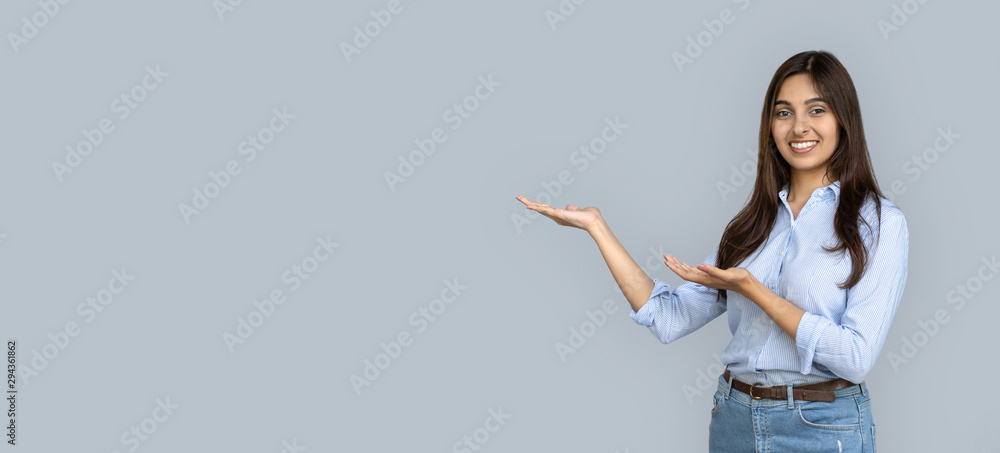 Smiling young adult indian woman stand isolated on grey background holding copy space on hands pointing. Happy girl student professional looking at camera advertise product service concept, banner - obrazy, fototapety, plakaty 